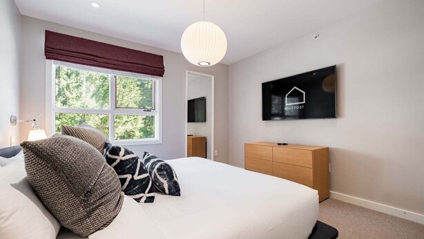 Ein Schlafzimmer mit einem Bett, einem an der Wand befestigten Fernseher, einem Fenster mit Blick auf Bäume und einer hölzernen Kommode.