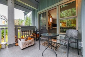 A covered patio area with a table and chairs, a wooden box labeled "Wood," and a large window with a view of trees. - Townhome | Complex hot tub | BBQ | Free Parking (Whistler)