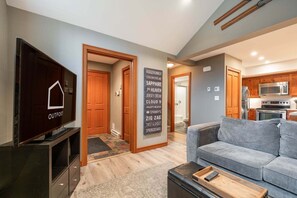 A modern living room with a grey sofa, a TV on a stand, a wooden door, and a kitchen visible in the background. - Townhome | Complex hot tub | BBQ | Free Parking (Whistler)