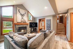A modern living room with a fireplace, a large television, and a stone wall with a deer painting. - Townhome | Complex hot tub | BBQ | Free Parking (Whistler)