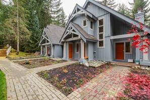 A two-story house with a steeply pitched roof, a front porch, and a well-maintained garden with a variety of plants and a small pond. - Townhome | Complex hot tub | BBQ | Free Parking (Whistler)