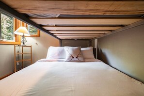 A single bed with white bedding and a unique pillow design, a bedside table with a lamp, and a window with a view of trees. - Townhome | Complex hot tub | BBQ | Free Parking (Whistler)