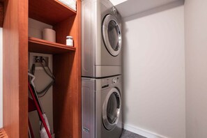 Ein Waschraum mit zwei übereinander gestapelten Waschmaschinen mit Frontlader, einem Holzregal mit einer Toilettenpapierrolle und einem Behälter, und einem roten Mop, der an der Wand lehnt.