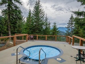 A hot tub surrounded by a wooden deck, overlooking a forested area with mountains in the distance. - Townhome | Wifi | Complex hot tub | BBQ (Whistler)