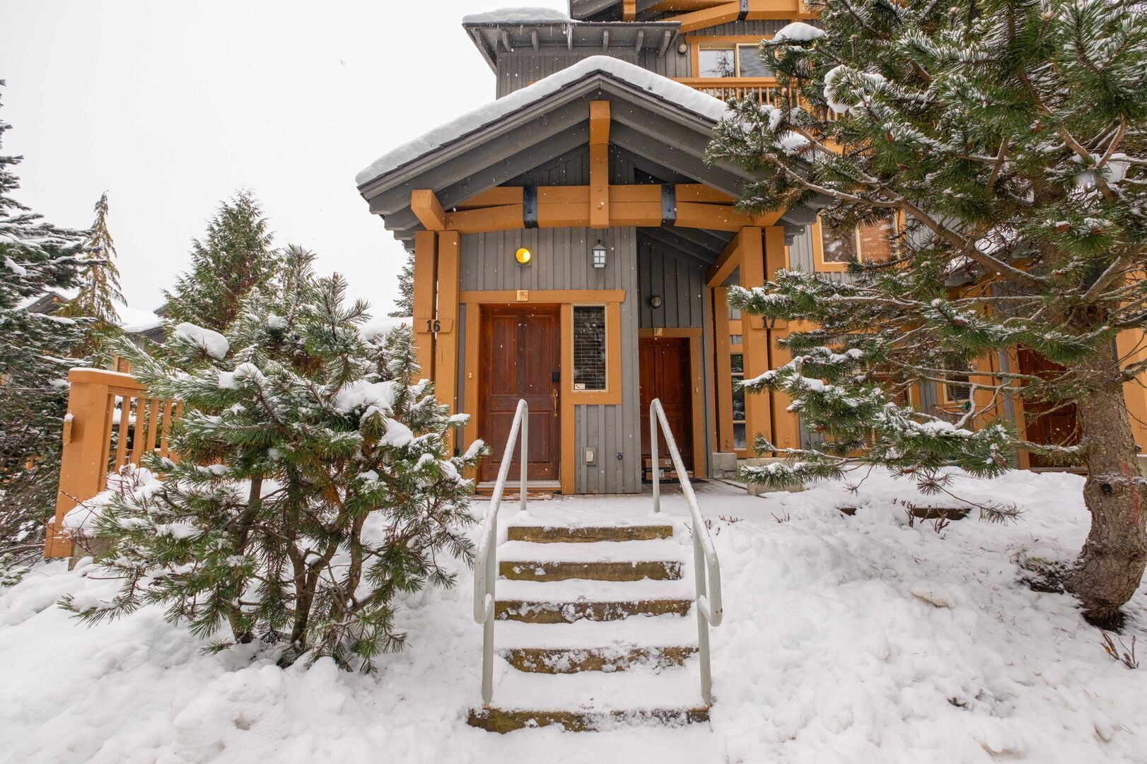 Un chalet en bois avec un escalier en bois menant à l’entrée, entouré d’arbres couverts de neige.