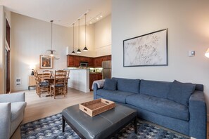 Ein modernes Wohnzimmer mit grauem Sofa, einem Essbereich mit Holzkästen und einer Küche mit dunklen Holzschränken sowie Pendelleuchten.
