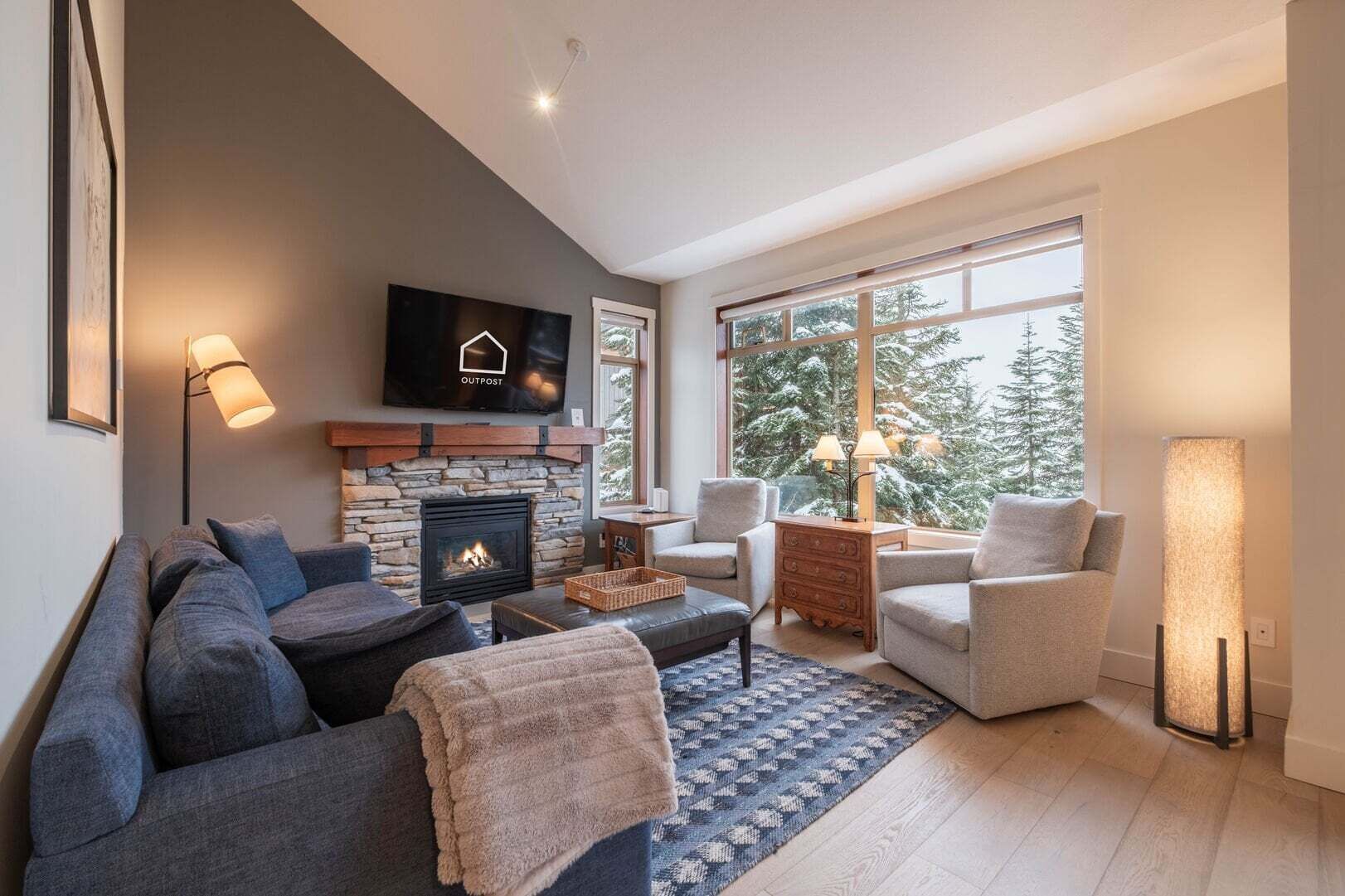 Ein modernes Wohnzimmer mit Kamin aus Stein, einem Flachbildfernseher und einem großen Fenster mit Blick auf schneebedeckte Bäume.