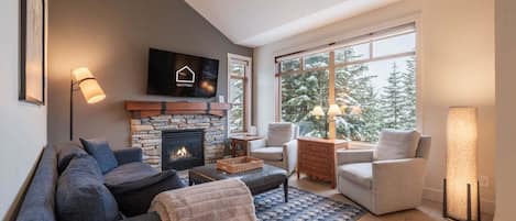 Een moderne woonkamer met een stenen open haard, een flatscreen televisie en een groot raam met uitzicht op met sneeuw bedekte bomen.