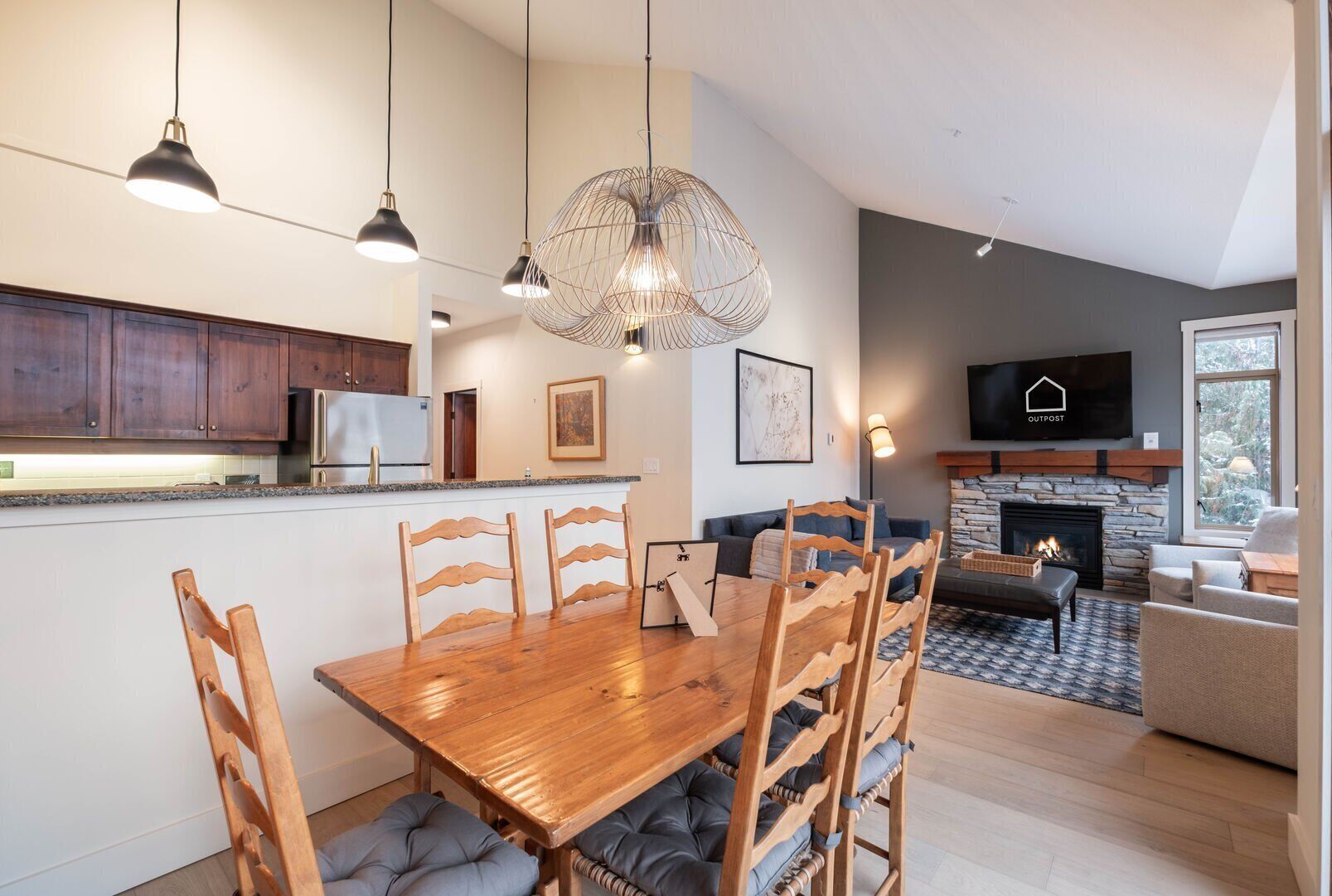 Una cocina moderna con armarios de madera y estufa integrada, un área de comedor con mesa y sillas de madera, y una sala de estar con chimenea y televisor montado en la pared.