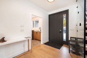 Ein modernes Interieur mit Holzboden, einer dunklen Tür, einer kleinen Küche mit Holzausstattung und einer weißen Wand mit Regal, auf dem eine kleine Statue steht.