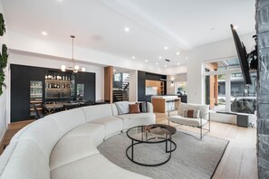A modern living room with a large L-shaped sectional sofa, a glass coffee table, and a kitchen area with dark cabinetry and a bar. - Gorgeous Chalet | Private hot tub | BBQ | WIFI (Whistler)