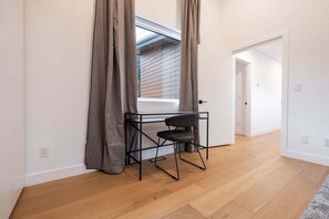 Een kamer met een bureau en stoel, een raam met gordijnen, houten vloeren, en een deur.
