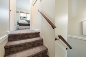 A carpeted staircase with wooden handrails, leading to a bedroom with a bed and a window. - Townhome | Private hot tub | Complex pool | BBQ (Whistler)