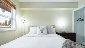 Ein Schlafzimmer mit Bett, Nachttisch, Lampe, Fenster und Blick auf Bäume.