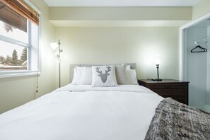 A bedroom with a bed, a bedside table, a lamp, a window, and a view of trees outside. - Townhome | Private hot tub | Complex pool | BBQ (Whistler)