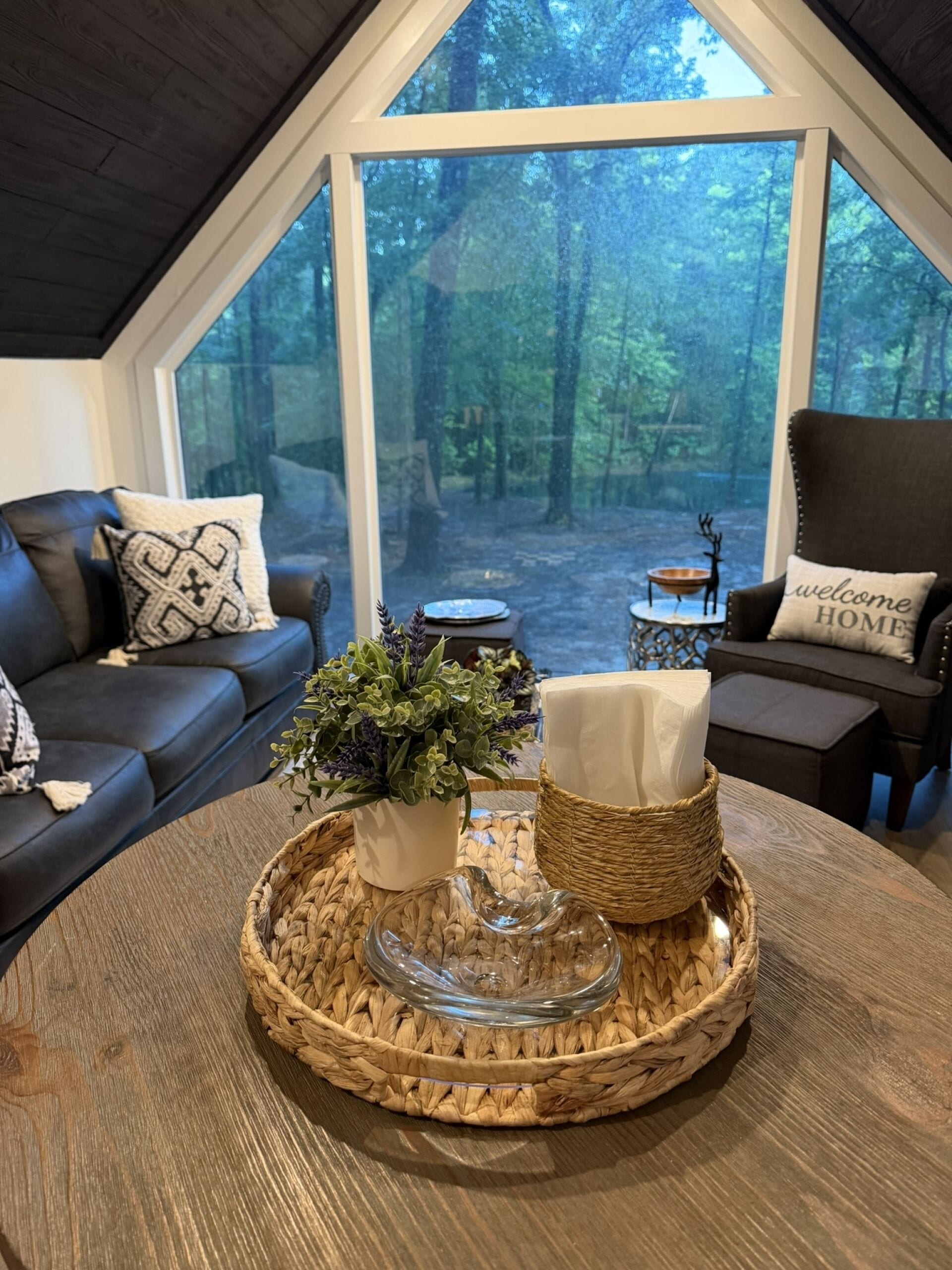 A cozy living room with a large window, a dark leather sofa, a round coffee table with a woven tray, and a wicker side table.