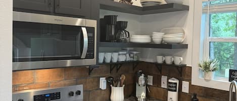 Een moderne keuken met een ingebouwde magnetron, fornuis en koffiezetapparaat. Boven de aanrecht zijn planken met servies en een kamerplant bij het raam.