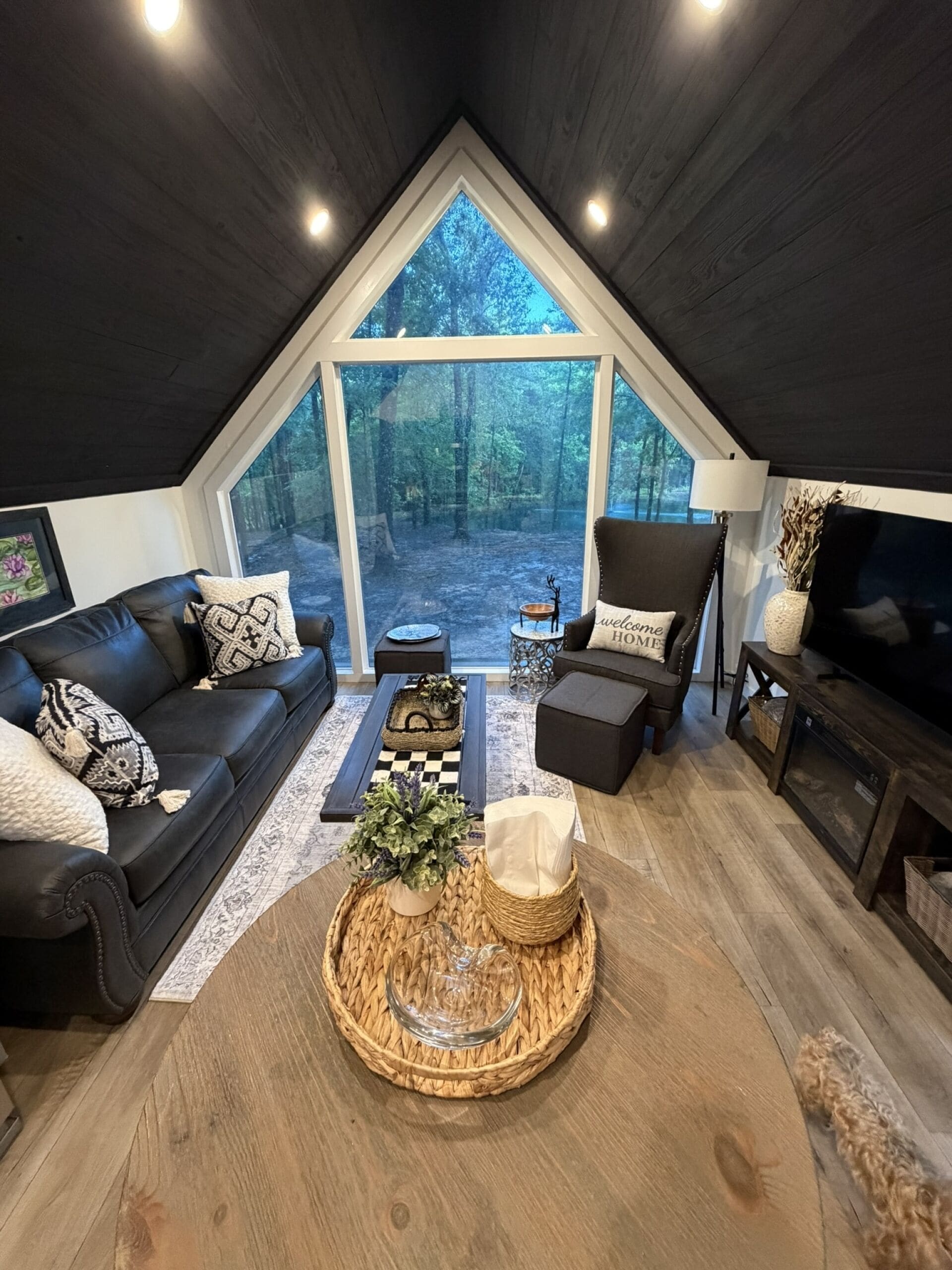 A cozy loft-style room with a large window, a black leather sofa, a wooden coffee table, and a TV on a wooden stand.