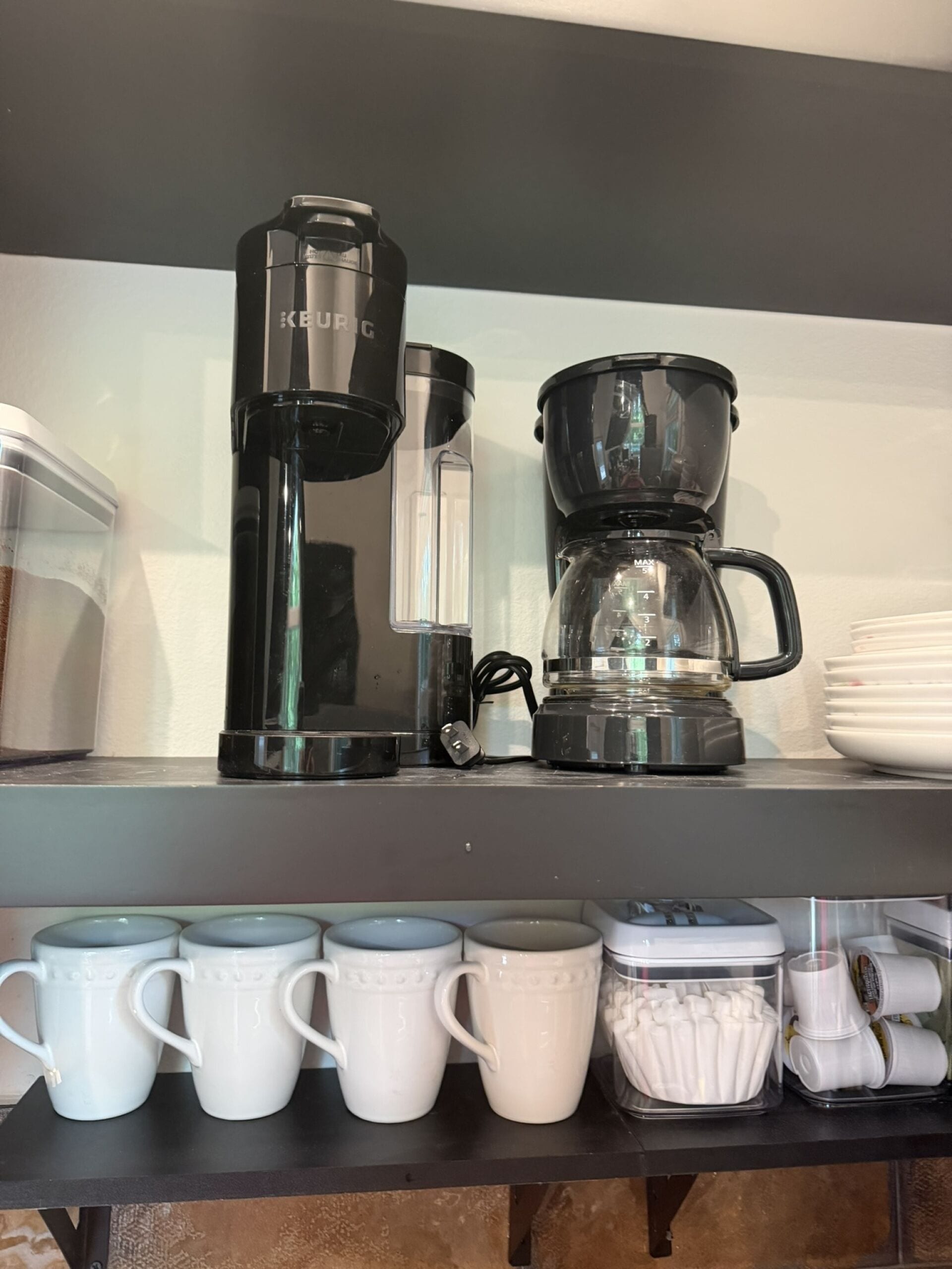 Un comptoir de cuisine avec une machine à café, une verseuse en verre et une cafetière. On y trouve des tasses blanches et un récipient contenant des ustensiles.