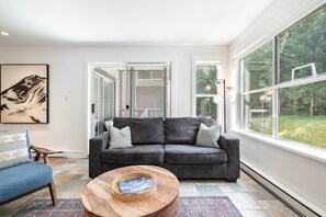 A living room with a grey sofa, a blue armchair, a wooden coffee table, and a large window offering a view of a garden. - Apartment | Complex hot tub | Free parking | Wifi (Whistler)