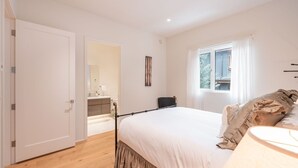 Quarto com uma cama grande, janela com cortinas, mesa de cabeceira e porta para um banheiro.