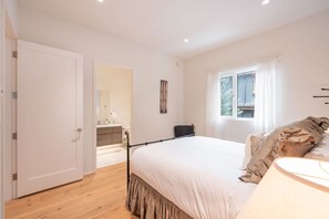 Ein Schlafzimmer mit einem großen Bett, einem Fenster mit Vorhängen, einem Nachttisch und einer Tür, die ins Badezimmer führt.