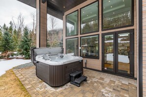 A modern outdoor hot tub area with a wooden deck, large windows, and a view of a snowy landscape. - Ultimate Secluded Chalet | Private hot tub (Whistler)