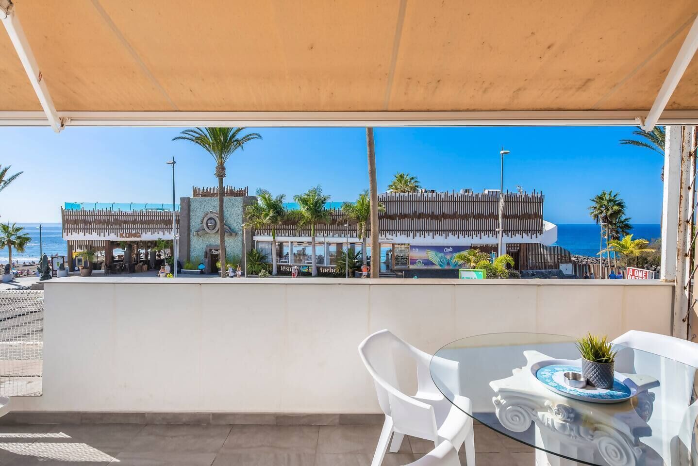 Ein Balkon mit einem Glastisch, weißen Stühlen und Blick auf Strand und Gebäude.