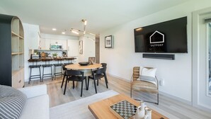 A modern living room with a dining area, a kitchen in the background, and a mounted flat-screen TV displaying 'OUTPOST'. - Ski-in/out | Complex pool, hot tub & BBQ | WIFI (Whistler)