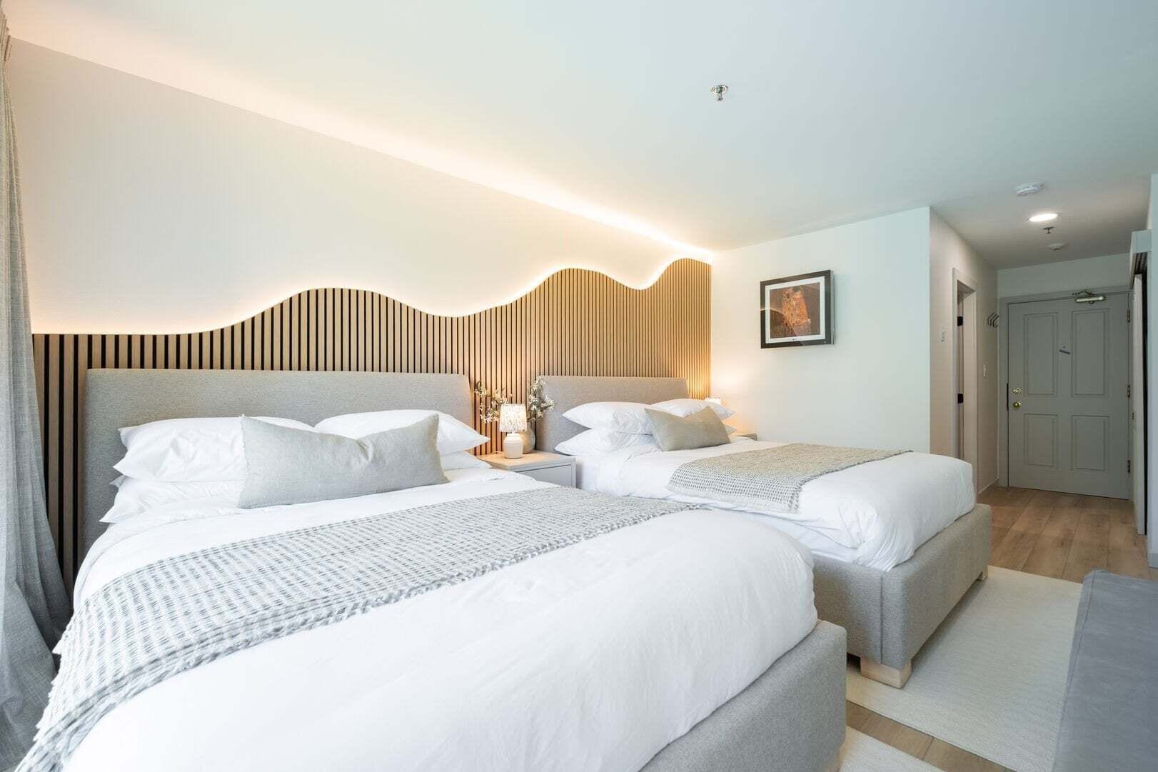 A hotel room with two beds, a wooden headboard, a bedside table with a lamp, and a framed picture on the wall.