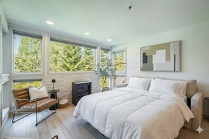 Une chambre à coucher avec un grand lit, un canapé, une cheminée et une vue sur les arbres par de grandes fenêtres.