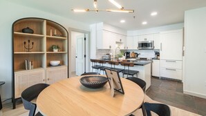 A modern kitchen with white cabinets, a central island, and a dining area with a round table and chairs. - Ski-in/out | Complex pool, hot tub & BBQ | WIFI (Whistler)
