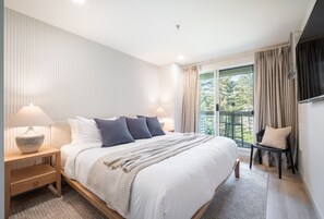 Une chambre moderne avec un grand lit, deux lampes de chevet, un fauteuil et une vue sur la verdure par la fenêtre.