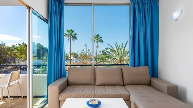 A room with a beige sectional sofa, a white coffee table, and a balcony with blue curtains. - 2 Bedroom Apartment with Seaview (Costa Adeje-Playas De Fañabe)
