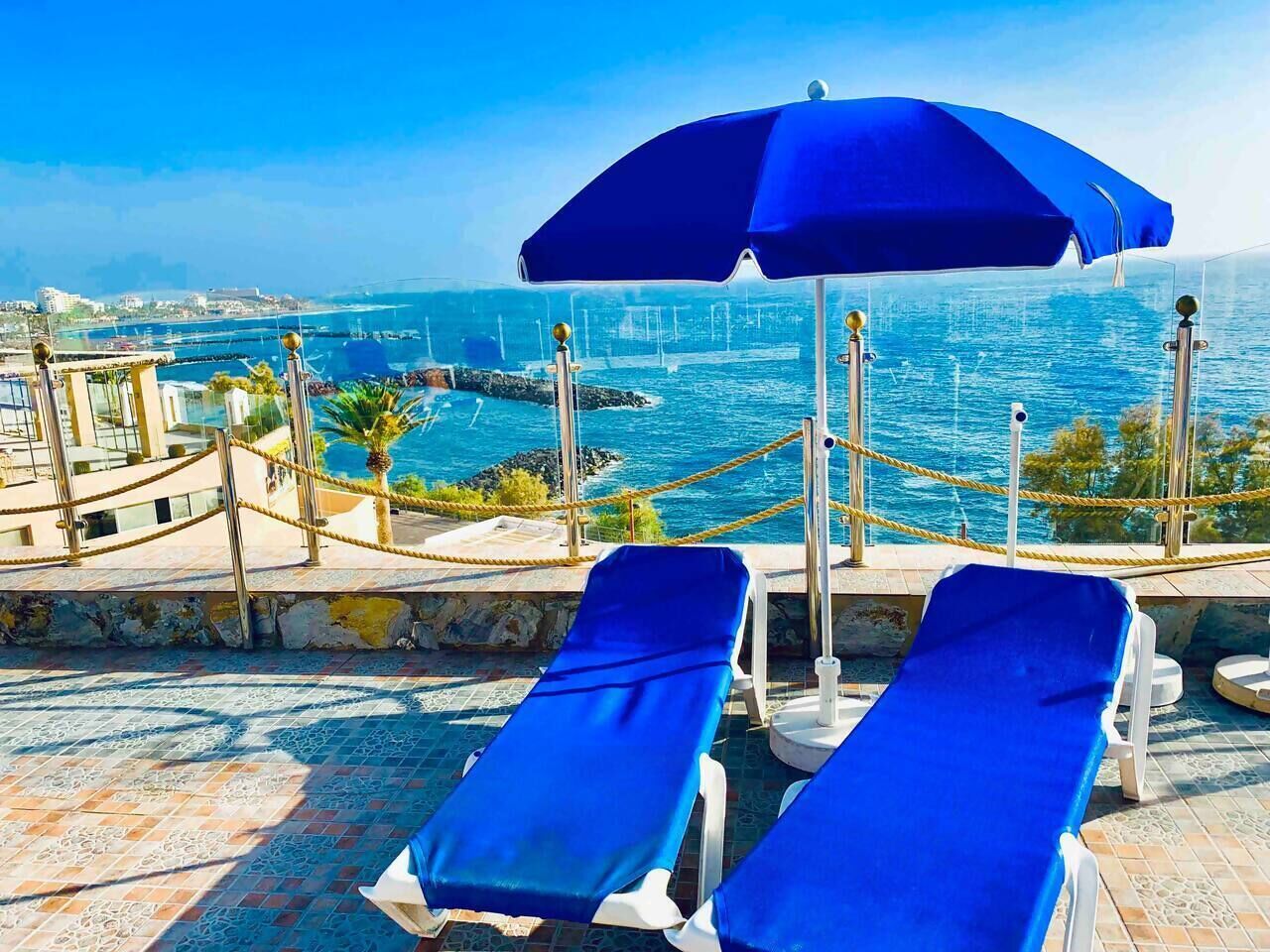 Zona de piscina con dos tumbonas azules y una sombrilla azul, con vistas a la costa.