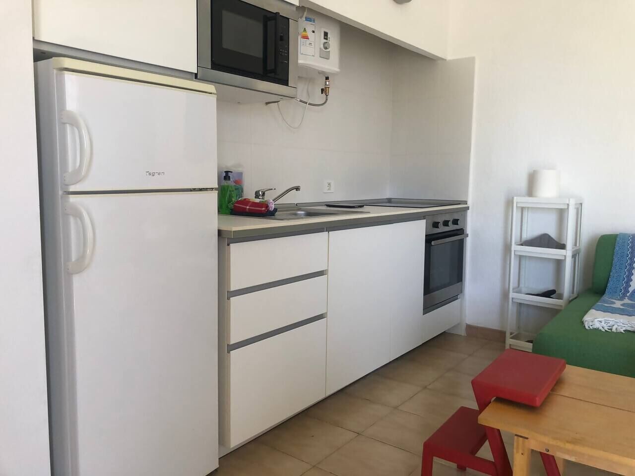 Cocina compacta con armarios blancos, un microondas, estufa y lavabo. En primer plano, hay una mesa pequeña y un taburete rojo.