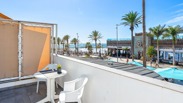 A balcony with a table and chairs, overlooking a beach with palm trees and a building with a clock. - Beachfront Studio Las Americas (Costa Adeje)