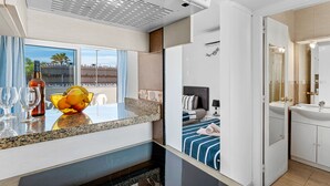 A modern hotel room with a kitchen area, a dining table with glasses, a fruit bowl, a bed with a blue and white striped bedspread, and a bathroom with a sink and mirror. - Beachfront Studio Las Americas (Costa Adeje)