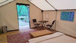 A tent with a bed, a table, two chairs, a trash bin, and a view of the forest. - Sorrel Tent | Canvas Tent with Lake Access (Kirkfield)