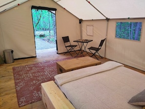 Una tenda con un letto, un tavolo, due sedie, un cestino e vista sulla foresta.