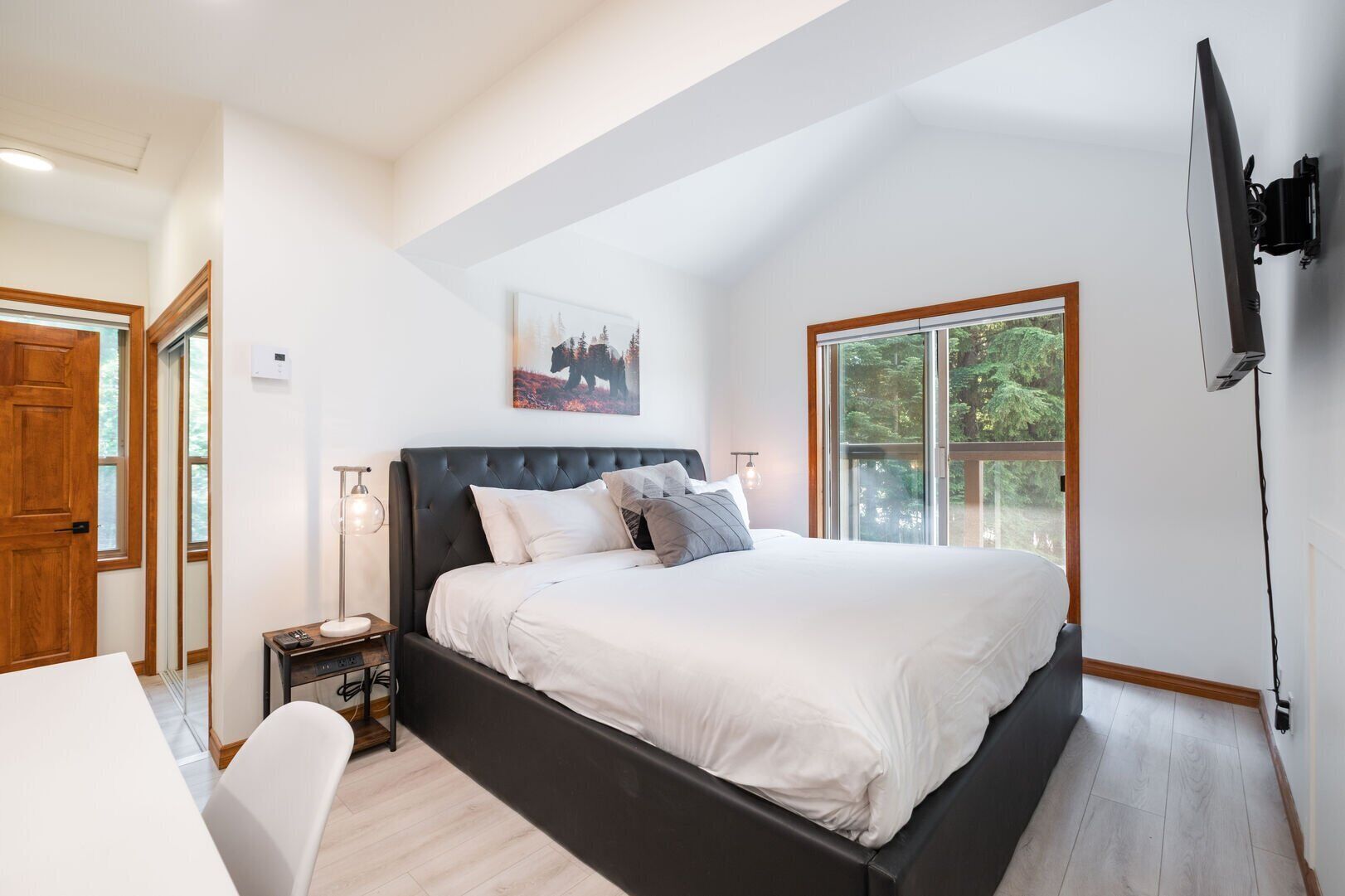 Une chambre avec un grand lit, une table de chevet, une lampe, un téléviseur fixé au mur et une fenêtre donnant sur des arbres.