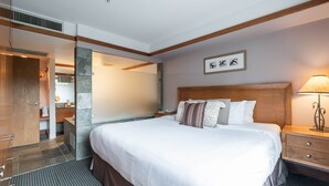 A hotel room with a large bed, wooden headboard, a nightstand, a lamp, and a bathroom visible through an open door. - Blackcomb Ski-in/out | Complex pool & hot tub (Whistler)