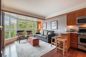 A modern living room with a large window, leather furniture, and a kitchen with wooden cabinets and stainless steel appliances. - Blackcomb Ski-in/out | Complex pool & hot tub (Whistler)