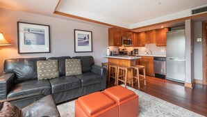 A modern living room with a leather sofa, a coffee table, and a kitchen area with wooden cabinets and stainless steel appliances. - Blackcomb Ski-in/out | Complex pool & hot tub (Whistler)