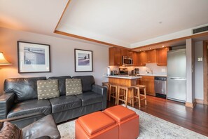 A modern living room with a leather sofa, a coffee table, and a kitchen area with wooden cabinets and stainless steel appliances. - Blackcomb Ski-in/out | Complex pool & hot tub (Whistler)
