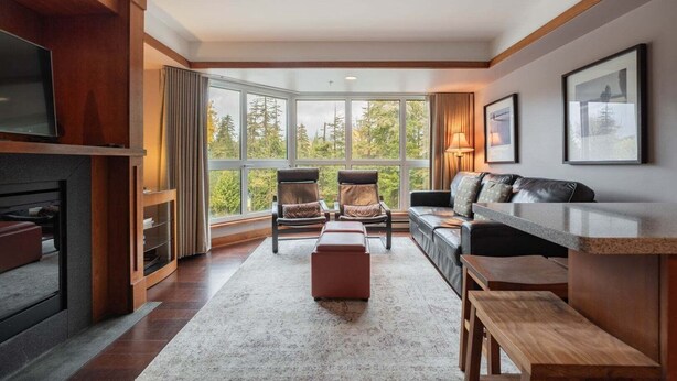 A modern living room with a fireplace, seating area, and large windows offering a view of trees. - Blackcomb Ski-in/out | Complex pool & hot tub (Whistler)