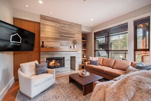 A modern living room with a fireplace, comfortable seating, and a large window offering a view of the outdoors. - Private Hot Tub | Central AC | BBQ | Garage (Whistler)