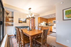 A dining area with a wooden table and chairs, a chandelier, and a view of the kitchen in the background. - Penthouse, Mountain Views, Ski-In/Out, Pool, Hot Tub, Gym (Whistler)