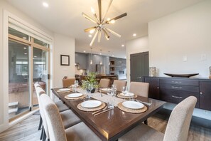 A modern dining room with a dark wooden table set for six, surrounded by beige chairs. A unique ceiling light fixture with multiple arms and bulbs is visible. - Luxury | Private garage | Private hot tub | BBQ (Whistler)