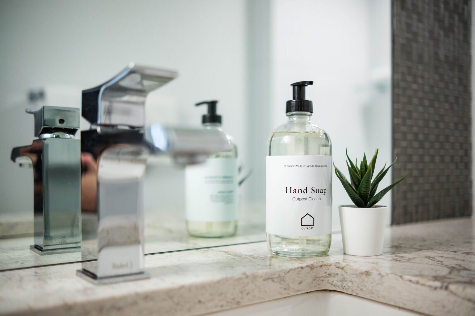 Uma pia de banheiro com uma torneira moderna, uma embalagem de sabonete líquido identificada como "Hand Soap Outpost Cleaner" e uma pequena planta em vaso sobre um balcão de mármore.